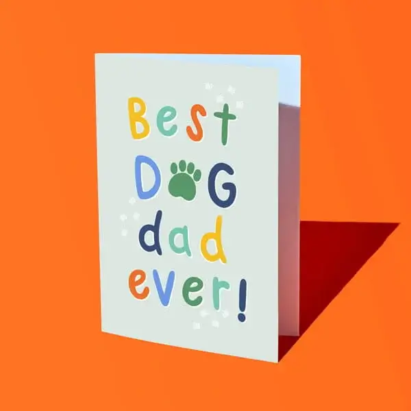 A Fathers Day card with colourful lettering reading Best Dog Dad Ever, featuring a paw print design, displayed against a bright orange background.
